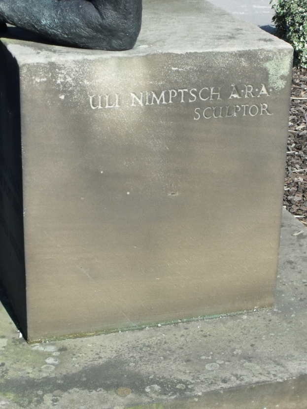 Ein Stein mit Text in Vordergrund, möglicherweise eine Statue oben, trockene Zweige, Erde und Pflanzen rechts, Straße im Hintergrund.
