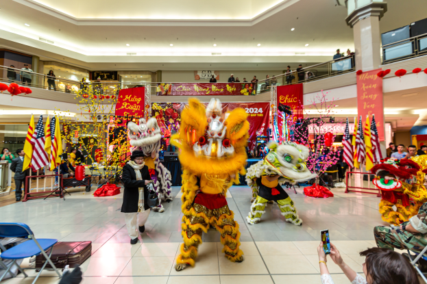 Lebendige Feier des chinesischen Neujahrs in einem Einkaufszentrum mit tanzenden Menschen in Kostümen und anderen auf Stühlen sitzend mit Mobiltelefonen, umgeben von Fahnen, Bannern, Dekorationen und Lichtern.