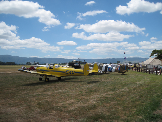 Flugzeuge, Fahrzeuge, Zelte und Fahnenmasten sind auf einer grünen Fläche mit Bäumen und Hügeln im Hintergrund unter einem klaren Himmel aufgebaut.