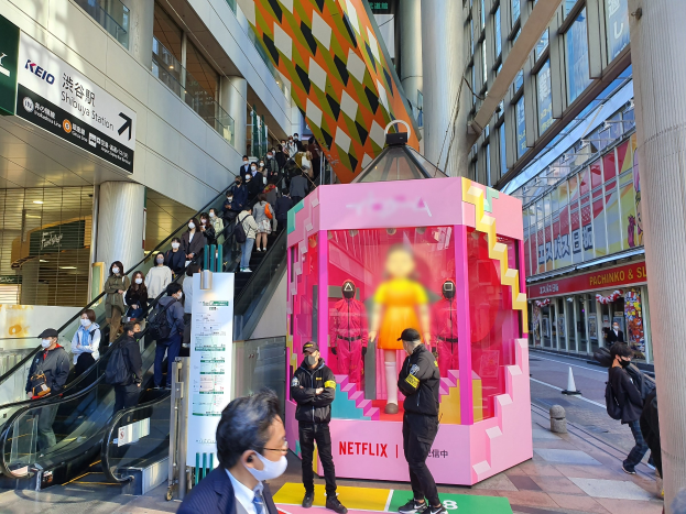 Eine Gruppe von Menschen steht vor einem Gebäude mit Glaswänden und Texttafeln, einige tragen Masken, während eine Rolltreppe mit Menschen daneben fährt; eine pinkfarbene Box in der Mitte zeigt Schaufensterpuppen und trägt den Text "Netflix's new Netflix booth in Tokyo."