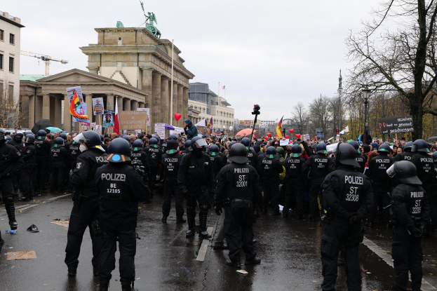 Eine große Gruppe von Polizisten steht vor einer Menge mit Schildern, Luftballons und Helmen, mit Gebäuden, einer Statue, einem Kran, Bäumen und einem bewölkten Himmel im Hintergrund.