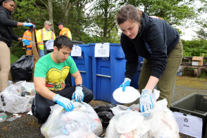 Eine Gruppe von Menschen, die Müll in einem Park sammeln, mit zwei Personen in der Mitte, die Handschuhe tragen und Schilder halten, umgeben von weggeworfenen Gegenständen, einem Mülleimer, einer Bank, Bäumen und einem klaren blauen Himmel.