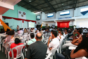 Eine Gruppe von Menschen sitzt auf Stühlen und schaut auf eine Bühne, auf der ein Polizeibeamter in ein Mikrofon spricht, wobei auf der rechten Seite des Bildes eine Tür zu sehen ist.
