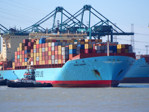 Ein große Container-Schiff liegt neben einem Schlepper, mit vielen Containern an Bord, und im Hintergrund gibt es Türme, Kabel und einen klaren blauen Himmel.