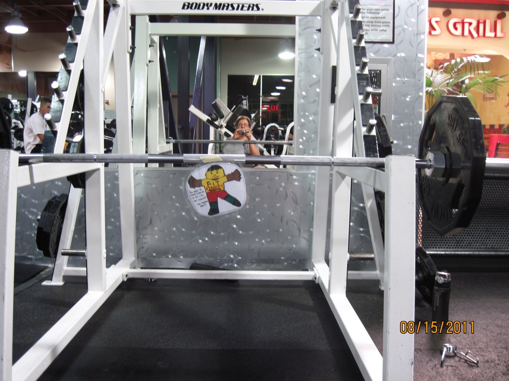 Ein Fitnessstudio mit einer Hantelbank im Vordergrund, einem Mann links und einer Frau in der Mitte sitzend.