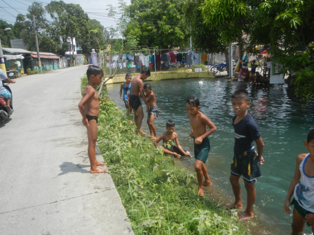 Kinder spielen in flachem Wasser neben einer Straße, einige stehen und andere sitzen auf Gras, mit einer Person auf einem Motorrad links und Häusern, Bäumen und einem klaren blauen Himmel im Hintergrund.