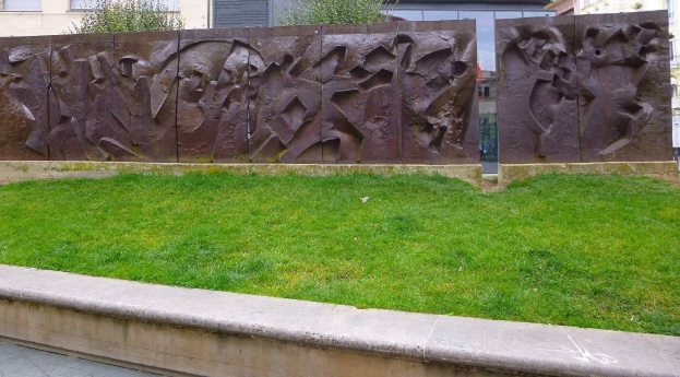 Eine große Metallskulptur des Berliner Walls steht auf einer grasbewachsenen Fläche umgeben von Bäumen und Gebäuden mit Fenstern im Hintergrund.
