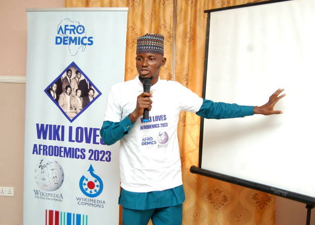 Ein Mann in einer Mütze hält ein Mikrofon und präsentiert vor einer Whiteboard, mit einem Banner und einem Vorhang im Hintergrund und einem Schaltpult an der Wand.