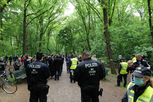 Polizeibeamte vor einer Menschenmenge bei einer Outdoor-Veranstaltung, mit Fahrrädern und einer Bank im Vordergrund, Bäumen und Himmel im Hintergrund.