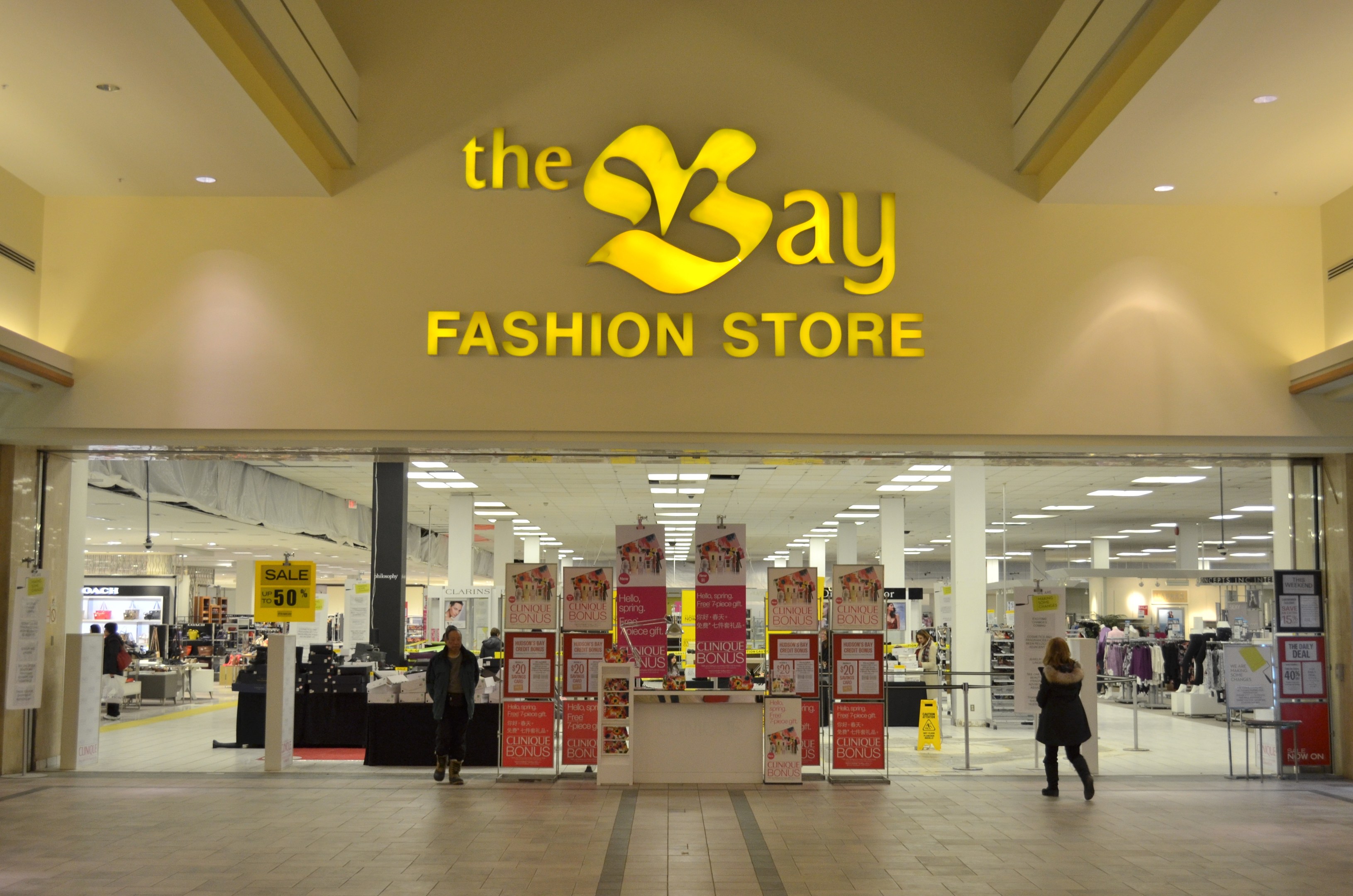 Außenansicht eines Bay Fashion Stores in einem Einkaufszentrum mit Menschen, Texttafeln an den Wänden, Säulen, Deckenleuchten und Hintergrundobjekten.