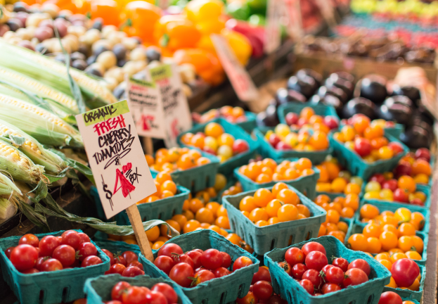 Ein Bauernmarkt mit Körben voller frischer Produkte wie Tomaten und Mais sowie Schilder mit Text im Hintergrund.