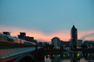 Ein Fluss mit weißen Booten, ein Zug auf einer Brücke rechts, Gebäude, Bäume, Lichter und Wolken im Hintergrund.