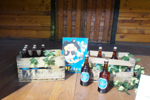 Ein Holzkasten voller Bierflaschen auf einem Holzboden, umgeben von Blättern und Blumen, mit einer Holzwand und einer Tür im Hintergrund.