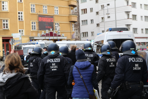 Eine Gruppe von Polizisten steht vor einer Menge von Menschen mit Helmen und Jacken während einer Demonstration in Berlin, Deutschland, mit Fahrzeugen, Gebäuden, Laternenpfählen und einem Banner mit Text im Hintergrund und einer Person mit einer Kamera.