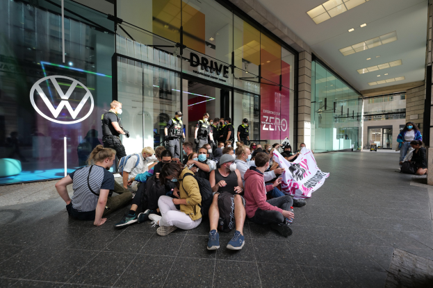 Eine Gruppe von Menschen sitzt vor einer Volkswagen-Vertretung mit Glaswänden auf dem Boden und hält eine Fahne mit Text, die zu protestieren scheint.