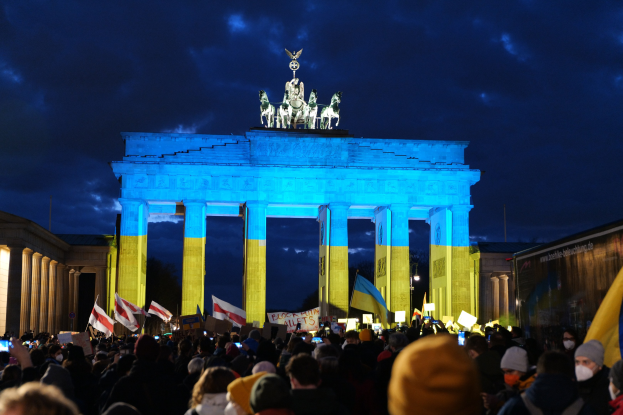 Eine Menschenmenge steht vor dem Brandenburger Tor in Berlin, Deutschland, mit Fahnen und Schildern in den Händen, einem Banner auf der rechten Seite und dem Tor mit Statuen und Säulen unter einem bewölkten Himmel.