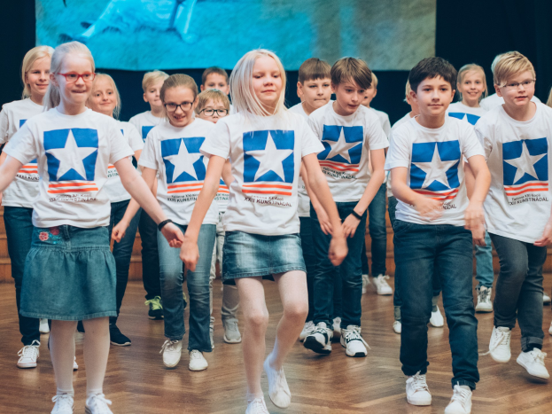 Gruppe von Kindern in weißen Hemden und blauen Shorts, die auf einem Holzfußboden vor einer Wand mit einem Bildschirm tanzen, an einem Kunstnädler-Tanzwettbewerb teilnehmen.