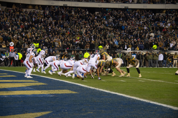 Ein Fussballspiel in einem Stadion mit Spielern in Sportkleidung und Helmen auf dem Feld, Zuschauern, Gelöndern und Tafeln mit Text im Hintergrund.