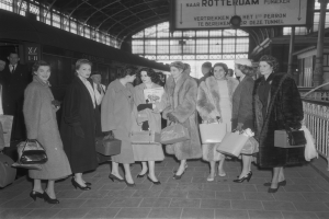 Schwarz-weißes Bild einer Gruppe von Frauen, die nebeneinander an einem Bahnhof stehen, jede hält eine Tasche in der Hand, mit einem Zug auf der linken Seite und Geländern und einer Tafel auf der rechten Seite.