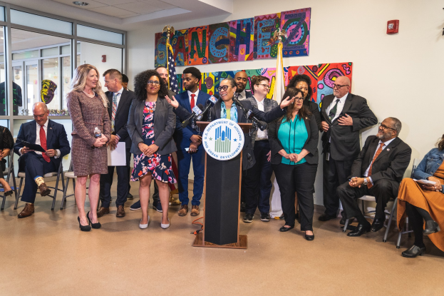 Eine Gruppe von Menschen, die in einem Raum mit einem Podium, einem Mikrofon und einer Tafel mit der Aufschrift "Sancho City Councilman Joins City Council" stehen und sitzen, mit Flaggen, Feuerlöscher und Deckenleuchten an der Rückwand.