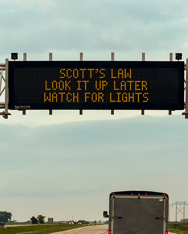 Ein Lkw fährt auf einer Autobahn mit einem Schild an der Seite, auf dem steht: "Scott's Law: Look it Up Later, Watch for Lights", umgeben von Gras, Bäumen und Pfählen, mit einem klaren Himmel im Hintergrund.