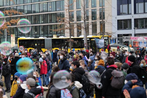 Große Gruppe von Menschen vor einem Gebäude mit Fenstern, Bäumen und Fahrzeugen während einer Demonstration in Berlin, einige halten Schilder und tragen Mützen oder Taschen, mit Blasen in der Luft.