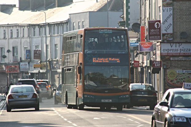 Eine Stra├če mit Autos und einem Bus im Vordergrund, mit Geb├Ąuden, die W├Ąnde, Fenster, T├Âpfe und D├Ącher im Hintergrund aufweisen, verziert mit Plakaten und Bannern, und einem Laternenmast.
