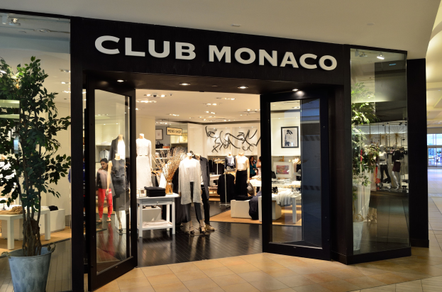 Vitrine eines Club Monaco-Geschäfts in einem Einkaufszentrum mit Glaswänden und -türen, Schaufensterpuppen in Kleidern, Auslagetischen mit Waren, einem Topf mit einer Pflanze und Deckenbeleuchtung.