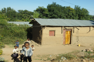 Kinder stehen neben einer H√ľtte mit einem Dach, Fenster und einer T√ľr mit gelben Vorh√§ngen, umgeben von B√§umen und √∂berirdischen Leitungen.