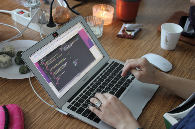 Ein Tisch mit einem Laptop, an dem eine Person tippt, eine Steckdose mit festen Kabeln, ein Teller mit Essen, eine Tasche, zwei Gl√§ser, eine Glaskerze, eine Verpackung, ein Becher und eine Maus.
