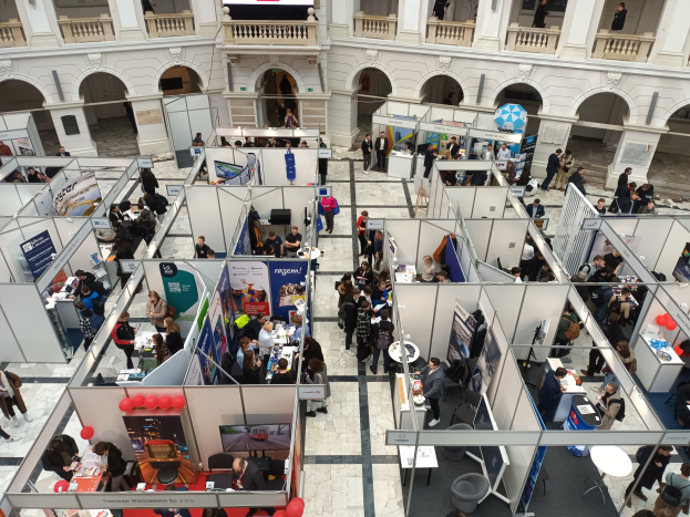 Eine große Gruppe von Menschen versammelt sich in einem Raum voller Stände, Banner, Tische, Stühle und Ballons, mit einem Gebäude mit Bögen, Säulen, Geländern und einem Bildschirm im Hintergrund.