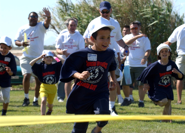 Kinder bei einem Lauf auf einer grünen Wiese mit einem gelben Band im Vordergrund, einige tragen Mützen und Schutzbrillen, andere halten Gegenstände, vor einem Hintergrund aus Bäumen und einem klaren blauen Himmel.