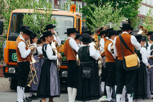 Gruppe von Menschen in traditioneller bayrischer Tracht, die eine Straße entlangmarschieren, Instrumente spielen und Taschen tragen, mit Bäumen und Gebäuden im Hintergrund und einem geparkten Fahrzeug.