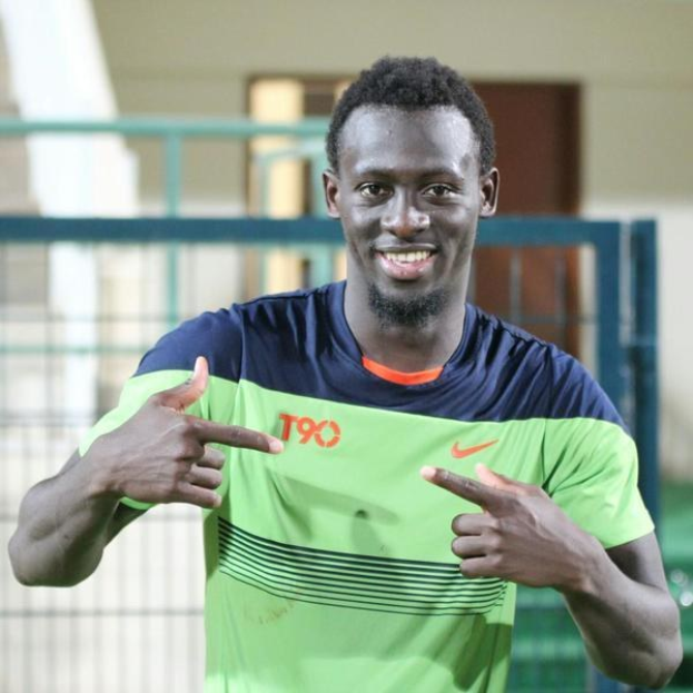 Ein Mann in einem grünen und blauen Hemd lächelt, während er auf die Kamera zeigt, mit einer unscharfen Reling, Wand und Tür im Hintergrund.