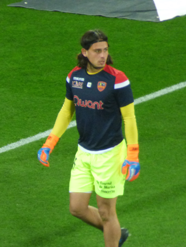 Ein Mann in einem blauen Hemd und gelben Shorts läuft auf einem Fußballfeld und streckt die Arme aus, mit einem entschlossenen Gesichtsausdruck und einem Tuch oben auf dem Bild.