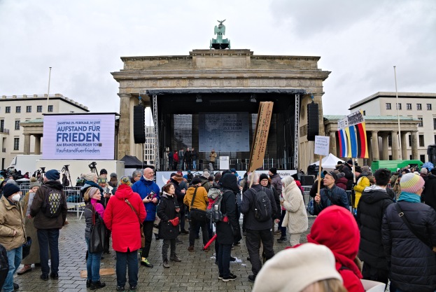 Eine Menschenmenge steht vor einem Gebäude mit einer Bühne, auf der Redner und ein Bildschirm zu sehen sind, sowie rechts davon Fahnen und Transparente, was auf eine Demonstration in Berlin hindeutet.