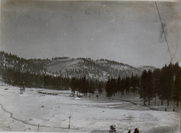 Ein Schwarz-Weiß-Foto einer Skipiste mit Menschen, die darauf Ski fahren, umgeben von Bäumen und Pfählen, mit Hügeln und einem klaren Himmel im Hintergrund und Text am unteren Rand des Bildes.