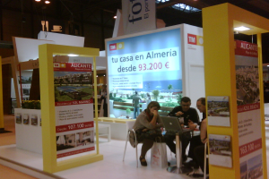 Eine Gruppe von Menschen, die um einen Tisch herum auf Stühlen sitzen und auf einen Laptop schauen, mit verschiedenen Werbeplakaten daneben.