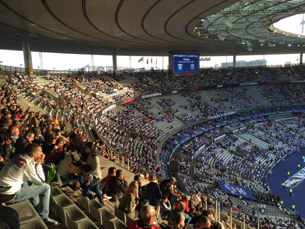 Große Menschenmenge in einem Stadion bei einem Fußballspiel, mit einer Bühne rechts, Fahnen, Stangen, einem Bildschirm und der Allianz Arena in München im Hintergrund.