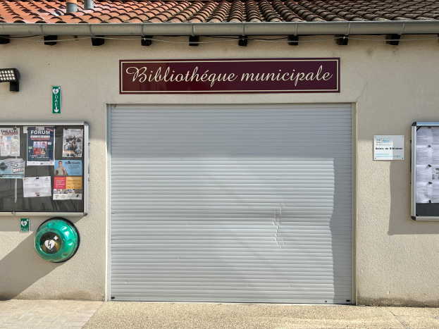 Ein Gebäude mit einer Tafel, auf der "Bibliothèque municipale" steht, mit einem Rollladen in der Mitte, einer Tafel mit Text, Plakaten an der linken Wand und einer Lampe, die den Bereich beleuchtet, mit einem Weg, der zur Tür führt.