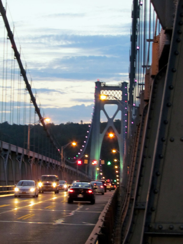 Fahrzeuge auf einer Brücke mit Lichtern, Masten, Hügeln und Himmel im Hintergrund.