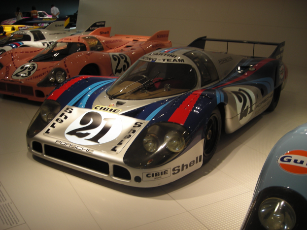 Ein Porsche 917K Rennwagen im Le Mans Museum, umgeben von anderen Sportwagen und Menschen, mit einer Wand und informativem Text im Hintergrund.