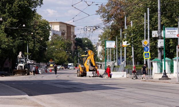 Stadtstraße mit einer Baustelle, Fahrzeugen, Fußgängern, einem Radfahrer, Verkehrskegeln, Pfählen, Schildern, Strommasten mit Drähten, Bäumen, Gebäuden mit Fenstern und einem bewölkten Himmel.