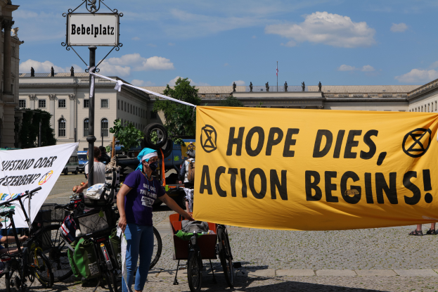 Eine Gruppe von Menschen steht vor einem Gebäude und hält ein gelbes Banner mit der Aufschrift "Hope Dies, Action Begins". In der Nähe stehen Fahrräder und es gibt einen Pfahl mit einer Tafel. Im Hintergrund sind Bäume und ein klarer blauer Himmel zu sehen.