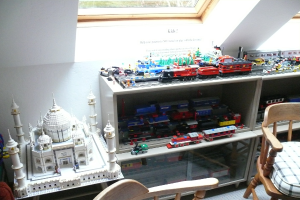 Ein Glasfenster oben in der Mitte, ein Stuhl rechts, eine Vitrine mit Spielzeugzügen in einer Reihe und ein Spielzeug Taj Mahal links auf einem Schreibtisch.