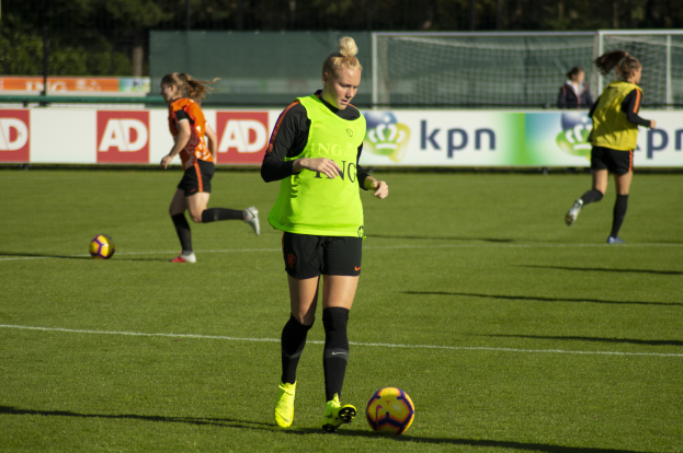 Eine Gruppe von Frauen beim Fußballspielen auf einem von Bäumen umgebenen Feld, mit Bannern im Hintergrund und einem Netz im Vordergrund.