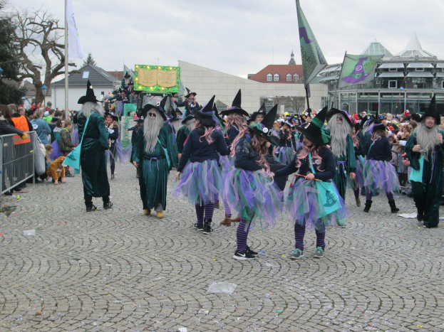 Eine Gruppe von Menschen in Hexenkostümen, die schwarze und lila Kleider und Hüte tragen und einige Fahnen halten, gehen eine Kopfsteinpflasterstraße entlang, neben einem Geländer mit Gebäuden, Bäumen und einem klaren blauen Himmel im Hintergrund.