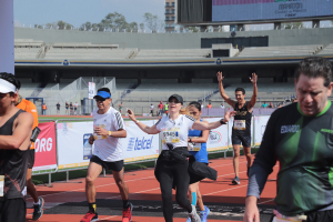 Gruppe von Läufern auf einer Außenbahn in einem Stadion mit Bannern, einem Display, einem Gebäude, Bäumen und einem bewölktem Himmel; einige Läufer tragen Turnschuhe und Hüte.