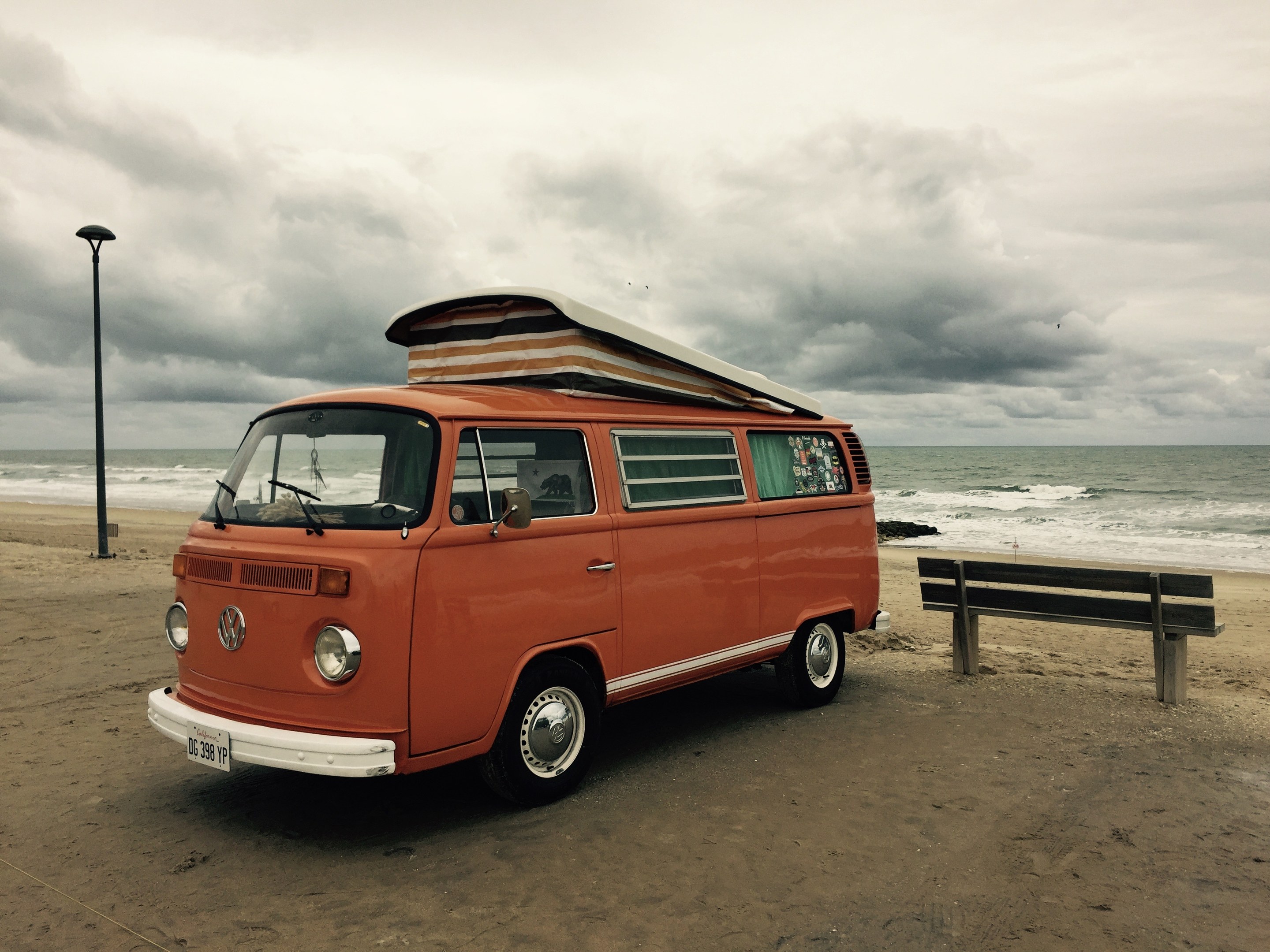 Oranger VW-Bus auf einem Strand neben dem Ozean geparkt, mit einer Bank und einem Laternenpfahl in der N├Ąhe, unter einem bew├Âlktem Himmel mit sichtbarem Wasser im Hintergrund.
