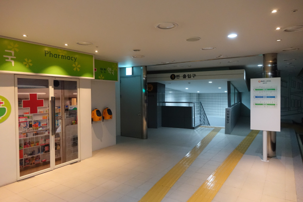 Apotheken-Eingang mit einem "Apotheke"-Schild auf der linken Seite, einer Tafel mit Text auf der rechten Seite, beleuchteten Lampen oben und Hintergrundobjekten.